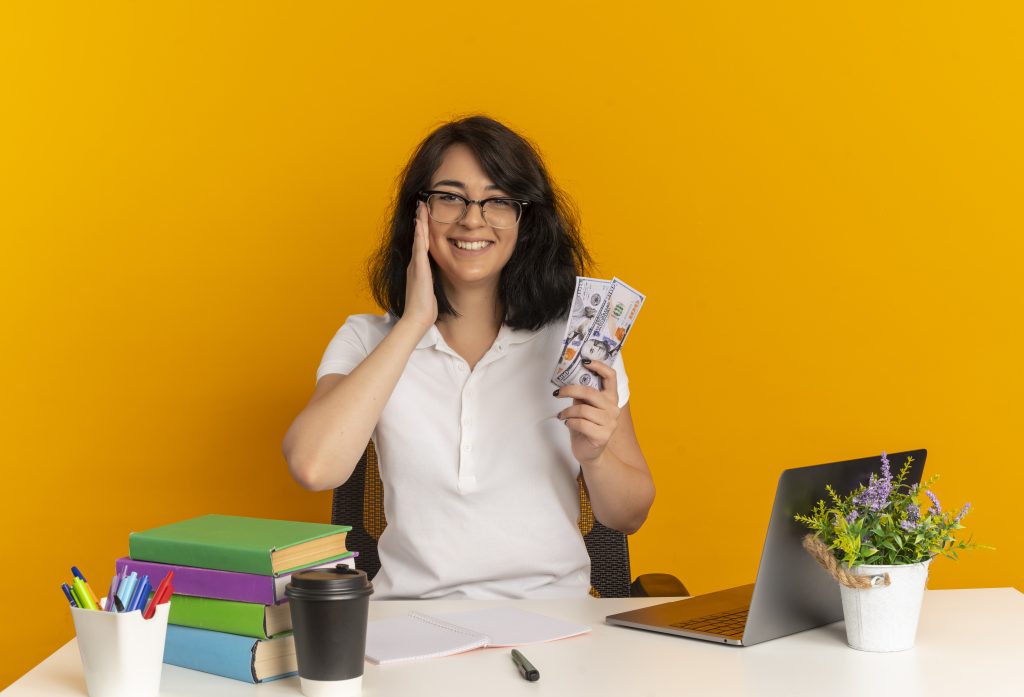 young smiling pretty caucasian schoolgirl wearing glasses sits at desk with school tools puts hand on face holds money isolated on orange background with copy space